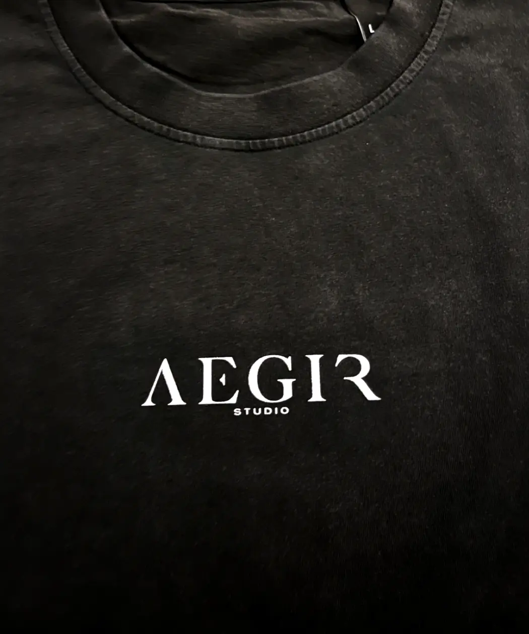 Close-up detail of the Aegir Studio x Jōhatsu tee, showing the texture of the vintage black organic cotton fabric and the crisp, white screen-printed 'AEGIR STUDIO' logo below the collar. - Close Up Thumbnail